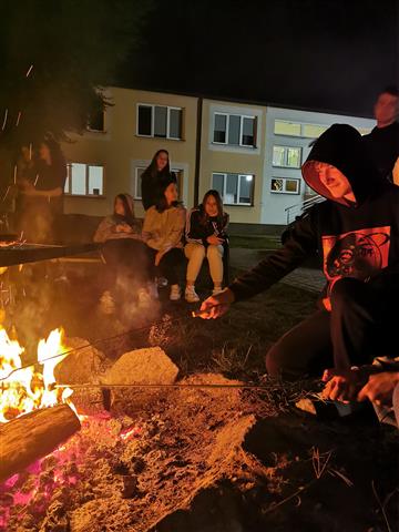 Uśmiechnięty wychowanek smaży kiełbaskę nad ogniskiem.  Na drugim planie wychowanki siedzą na ławce. W tle widać budynek bursy. 