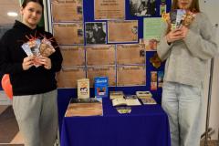 Dwie wychowanki stoją przy stoliku, trzymając zakładki do książek pt. Patron roku 2025  na Mazowszu – Antoni Słonimski. Na stoliku znajdują się książki Antoniego Słonimskiego. Za wychowankami znajduje się wystawa: biografia i zdjęcia Antoniego Słonimskiego.