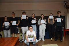 Siedmioro wychowanków stoi, jeden wychowanek kuca przed wszystkimi. Wychowankowie trzymają w rękach kartki z napisami, wyrażanie różnych emocji i gdzie można zwrócić się o pomoc. Numer telefonu zaufania dla dzieci i młodzieży 116 111