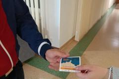 Jedna osoba wręcza drugiej osobie na korytarzu bursy ulotkę edukacyjną z numerem telefonu zaufania dla dzieci i młodzieży