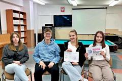 Czwórka wychowanków siedzi na krzesłach, dwie osoby trzymają materiały związane z „Dniem Bezpiecznego Internetu”