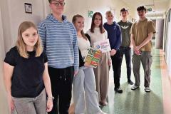 Młodzież stoi w korytarzu, trzymając materiały edukacyjne o zagrożeniach w internecie. Wychowankowie są uśmiechnięci, a otoczenie tworzy spokojną, domową atmosferę.
