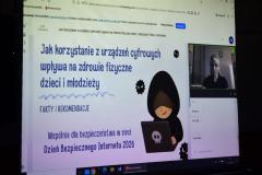 Na ścianie wyświetlana jest strona tytułowa webinaru nt. „Jak korzystanie z urządzeń cyfrowych wpływa na zdrowie fizyczne młodzieży? Fakty i rekomendacje. Wspólnie dla bezpieczeństwa w sieci. Dzień Bezpiecznego Internetu 2026 r.