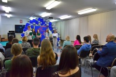 Sala wypełniona publicznością skierowaną w stronę grupy pięciorga wychowanków stojących na scenie, którzy prezentują program artystyczny z okazji Dnia Edukacji Narodowej. W tle widoczna jest dekoracja z balonów w odcieniach niebieskiego i srebrnego oraz tablica z napisem 'Dzień Edukacji Narodowej'.