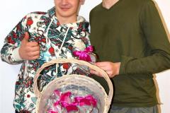 Dwóch chłopców prezentuje zawartość wiklinowego koszyka – słodkie upominki w kształcie serca zapakowane w folię i przewiązane różowymi kokardami.