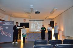 Młodzież w skupieniu prowadzi wieczór patriotyczny, w tle tablica z napisem 11 listopada, Narodowe Święto Niepodległości, Białym Orłem i liśćmi w kolorze biało czerwonym oraz dekoracja z liśćmi jesiennymi , wśród których stoją zapalone lampiony.