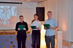 Młodzież w skupieniu prowadzi wieczór patriotyczny, w tle dekoracja z liśćmi jesiennymi , wśród których stoją zapalone lampiony.