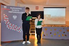 Młodzież w skupieniu prowadzi wieczór patriotyczny, w tle tablica z napisem 11 listopada, Narodowe Święto Niepodległości, Białym Orłem i liśćmi w kolorze biało czerwonym.