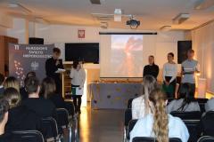 Młodzież w skupieniu prowadzi wieczór patriotyczny, w tle tablica z napisem 11 listopada, Narodowe Święto Niepodległości, Białym Orłem i liśćmi w kolorze biało czerwonym oraz dekoracja z liśćmi jesiennymi , wśród których stoją zapalone lampiony. Przed występującymi siedzi młodzież i wychowawcy. – zdjęcie jako główne