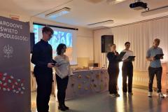 Młodzież w skupieniu prowadzi wieczór patriotyczny, w tle tablica z napisem 11 listopada, Narodowe Święto Niepodległości, Białym Orłem i liśćmi w kolorze biało czerwonym oraz dekoracja z liśćmi jesiennymi , wśród których stoją zapalone lampiony.