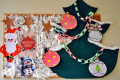 Widoczna świąteczna gazetka, na której pięknie prezentuje się zielona choinka udekorowana kolorowymi papierowymi bombkami i łańcuchem. Obok, na białej wacie, znajduje się Mikołaj z bałwankiem, a całość dopełniają świąteczne życzenia z napisem „Wesołych Świąt”