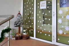 pięknie udekorowany kącik na kondygnacji przygotowany przez wychowanków. Widać choinkę wykonaną z waty oraz świąteczne pudełka ozdobione papierem dekoracyjnym i kokardami. Na jednym z nich stoi biały bałwanek, który dodaje całości uroku i świątecznego klimatu. Obok, na szybach, umieszczono białe papierowe gwiazdki oraz śnieg, które dodatkowo podkreślają zimową atmosferę