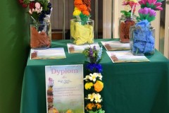 Stolik przykryty zielonym płótnem. Przed stolikiem stoi różnokolorowa plama wielkanocna. Z boku przypięty jest dyplom za zajęcie III miejsca w III międzyinternackim konkursie plastycznym na „Palmę Wielkanocną”. Na stoliku w szklane słoiki włożone są cztery palmy wielkanocne (kurpiowskie) oraz dyplomy za udział w międzyinternackim konkursie plastycznym na „Palmę Wielkanocną”.