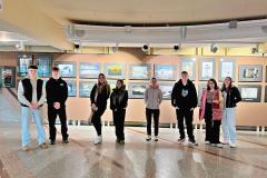 Grupa młodzieży stoi w korytarzu, w tle znajdują się ściany z fotografiami. Młodzi ludzie ubrani są w codzienne stroje, a ich postawa jest swobodna. Oświetlenie podkreśla ekspozycję prac artystycznych, które przedstawiają pejzaże, portrety oraz inne kreatywne ujęcia.