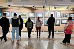 Grupa młodzieży ogląda wystawę fotografii i rysunków na korytarzu wystawowym. Osoby stoją tyłem do obiektywu, skupiając uwagę na prezentowanych pracach, które przedstawiają krajobrazy, portrety i artystyczne kompozycje. Oświetlenie podkreśla szczegóły eksponatów, tworząc klimatyczną atmosferę.