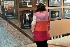 Wychowanka ogląda wystawę fotografii i rysunków na korytarzu wystawowym. Stoi tyłem do obiektywu, skupiając uwagę na prezentowanych pracach, które przedstawiają krajobrazy, portrety i artystyczne kompozycje. Oświetlenie podkreśla szczegóły eksponatów, tworząc klimatyczną atmosferę.