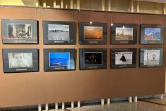 Fragment wystawy fotograficznej prezentującej dziesięć zdjęć w eleganckich, czarnych ramkach, zawieszonych na brązowej ścianie. Prace przedstawiają różnorodne motywy, w tym pejzaże, architekturę, portrety oraz artystyczne kompozycje. Zdjęcia są starannie oświetlone, podkreślając ich detale i klimat.