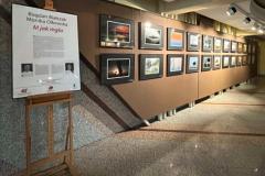 Fragment wystawy fotograficznej prezentującej zdjęcia w eleganckich, czarnych ramkach, zawieszonych na brązowej ścianie. Prace przedstawiają pejzaże. Zdjęcia są starannie oświetlone, podkreślając ich detale i klimat. Na sztaludze stoi tablica informacyjna.