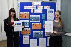 Dwie wychowanki bursy stoją przy niebieskiej tablicy na której znajduje się wystawa z okazji Ogólnopolskiego Dnia Walki z Depresję. W rękach trzymają materiały edukacyjne nt. Antydepresyjny Telefon oraz Jak rozmawiać z chorym na depresję?.