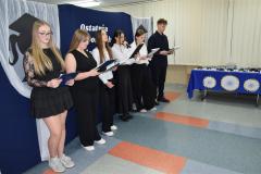 Wychowankowie stoją na tle dekoracji. Z boku znajduje się stół z nagrodami i dyplomami.