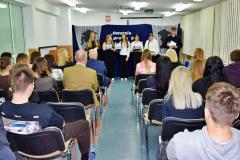 Grupa młodzieży stoi na scenie podczas uroczystego pożegnania maturzystów, a przed nimi siedzi publiczność.