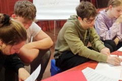 Wychowankowie pracują w grupach. Na stole leżą materiały potrzebne do zajęć. W tle znajduje się tablica z tematem spotkania.