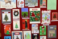 Czerwona na tablica na której znajdują się prace konkursowe (27 kart świątecznych przedstawiających: choinki, życzenia świąteczne, zimowe krajobrazy, choinki, ozdoby świąteczne). Na tablicy widnieje napis: XIII Międzyinternacki  konkurs plastyczny na kartkę bożonarodzeniową. 