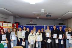 Na zdjęciu znajdują uczestnicy XIII Międzyinternackiego konkursu plastycznego na „Kartkę Bożonarodzeniową” wraz z opiekunami. W dłoniach trzymają dyplomy i nagrody. Z tyłu znajduje się czerwona tablica z kartkami konkursowymi i niebieska tablica z migającymi światełkami  i białymi gwiazdkami.