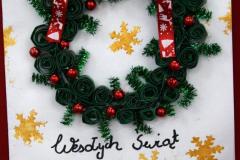 Kartka świąteczna: na białym tle znajduje się wieniec bożonarodzeniowy z zawiniętych w kółeczka zielonych pasków papieru, przeplatany czerwonymi perełkami. Na górze wieńca zawiązana jest czerwona kokardka w białe renifery i gwiazdki. Na kartce na białym tle znajdują się również złote gwiazdki. Na dole widnieje napis: Wesołych świąt. Pod kartką znajduje się napis: Wyróżnienie: Koziatek Amelia – Internat przy ZSZ nr 2 w Ostrołęce