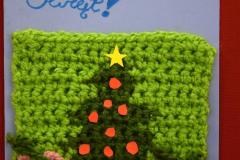 Kartka świąteczna: Na niebieskim tle znajduje się w formie kwadratu wydziergana na włóczce: ciemnozielona choinka na której przyczepione są czerwone bombki. Po bokach znajdują się różowe pudełka prezentowe zawiązane zieloną włóczką. Na górze znajduje się napis: Wesołych świąt. Pod kartką znajduje się napis: Wyróżnienie: Brzozowska Oliwia – Internat I LO im. gen. Józefa Bema w Ostrołęce