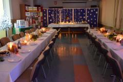 Sala w której  na pierwszym planie znajdują się dwa długie stoły z krzesłami : przykryte białymi obrusami. Na stołach znajdują się świece w szklanych wazonach, talerze, szklanki, dzbanki z wodą i sokami. Na talerzach znajdują się owoce i ciastka. Po lewej stronie znajduje się czerwona tablica na której znajdują prace konkursowe (kartki świąteczne). Z boku stoi stolik  z nagrodami. Z tyłu stoi niebieska tablica z białymi gwiazdkami. Na tablicy wiszą zapalone lampki. Przed tablicą stoi stół przykryty białymi obrusami.  Na stole znajdują się dekoracje świąteczne: lamki, choinka, gałązki świerku. W sali panuje świąteczna i nastrojowa atmosfera. 