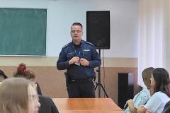 Policjant stoi i mówi do młodzieży siedzącej przy stolikach.