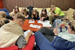 Grupa młodych osób bierze udział w zajęciach edukacyjnych. Wychowankowie siedzą wokół stołu na kanapach i z zaangażowaniem rozwiązują karty pracy 