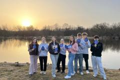 Grupa młodych osób pozuje do zdjęcia na tle wody i zachodu słońca. W rękach trzymają materiały promocyjne z akcji: Światowy Dzień Wody.