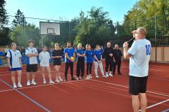 Uczestnicy Turnieju Piłki Siatkowej wysłuchują zasad rozgrywki. Pan Krzysztof Prot wita wszystkich zawodników. W tle kibice, siedzący na trybunie boiska.