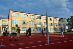 Akcja na boisku do siatkówki. Grupa osób gra w siatkówkę. W tle budynek bursy.