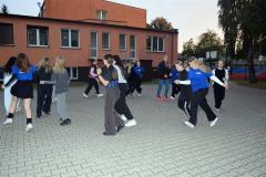Wychowankowie tańczą w parach i śmieją się. ”. Tańczą w rytm muzyki na świeżym powietrzu. W tle budynek bursy.