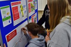 Grupa młodych osób pracuje przy wystawie o migracjach. Jedna osoba klęczy i zapisuje swoje refleksje na plakacie „cichej dyskusji”, a pozostali stoją obok i przyglądają się materiałom na tablicy.