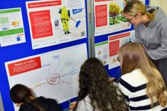 Kilka osób klęczy przy tablicy z wystawą o migracjach i dopisuje swoje refleksje na plakacie. Obok nich znajdują się materiały edukacyjne i plakaty dotyczące polskości oraz doświadczeń migracyjnych.