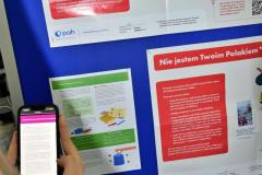 Wychowanka trzyma telefon i skanuje kod QR przy tablicy z plakatami PAH dotyczącymi migracji i polskości, przeglądając dodatkowe materiały na ekranie.