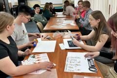 Grupa młodzieży uczestniczy w zajęciach edukacyjnych w ramach akcji „Edukujesz – Ratujesz”. Uczestnicy siedzą przy długim stole, pracując w parach lub małych grupach. Przed nimi znajdują się różne materiały dydaktyczne, takie jak ulotki, plakaty, arkusze papieru, flamastry, klej. Młodzież skupiona jest na przygotowywaniu materiałów związanych z pierwszą pomocą-niektórzy czytają instrukcje, inni wycinają elementy lub komponują plakaty. 