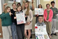 Grupa młodzieży prezentuje wykonane przez siebie plakaty edukacyjne w ramach akcji „Edukujesz – Ratujesz”. Wychowankowie stoją i klęczą w rzędach, trzymając przed sobą kolorowe prace dotyczące pierwszej pomocy – od opatrywania ran, przez złamania, aż po numery alarmowe. Uśmiechnięci i zaangażowani, pokazują efekty swojej kreatywności i zdobytej wiedzy.