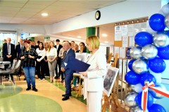 Pani Dyrektor odczytuje przemówienie do zgromadzonych gości oraz wychowanków, na holu głównym. Po prawej stronie widoczne niebieskie, srebrne i białe balony oraz biało czerwona wstęga.