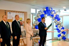 Uczestnicy uroczystości otwarcia stołówki stoją przy łuku z balonów w kolorach niebieskim i srebrnym. Uczestnicy uroczystości stoją obok wstęgi, a jedna z osób odczytuje dokument.