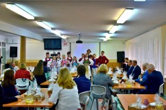 Grupa dzieci i młodzieży w tradycyjnych strojach ludowych, wykonuje występ artystyczny przed zgromadzoną publicznością. Widzowie, siedzący przy stołach, obserwują występ, a na stole widoczne są zapakowane upominki.