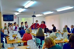 Grupa dzieci ubranych w stroje ludowe wykonuje tradycyjny taniec przed publicznością. Obok sceny widoczny jest muzyk grający na keyboardzie, a widzowie siedzą przy stołach.