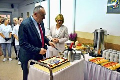 Mirosław Krusiewicz, dyrektor Departamentu Edukacji Publicznej i Sportu Samorządu Województwa Mazowieckiego, kroi okolicznościowy tort w towarzystwie dyrektor bursy regionalnej w Ostrołęce, Aliny Ambroziak. W tle znajdują się pracownicy stołówki ubrani w fartuchy, a na stole przygotowane przekąski i napoje.