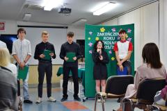 Grupa pięciorga uśmiechniętych wychowanków stoi na środku sali i prowadzi część artystyczną. W tle widać zieloną tablicę dekoracyjną z kwiatami i napisem „ZAKOŃCZENIE ROKU SZKOLNEGO 2025/2026”. Z prawej strony tyłem do obiektywu siedzi publiczność.