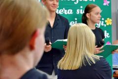 Zbliżenie na dwoje wychowanków (chłopaka i dziewczynę) trzymających mikrofony. Stoją uśmiechnięci na tle zielonej tablicy z białym napisem zaczynającym się od słów „Najważniejsze, gdy...”. Na pierwszym planie widać rozmyte sylwetki siedzącej publiczności.