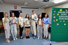 Uśmiechnięta grupa nagrodzonej młodzieży pozuje do zdjęcia z kadrą pedagogiczną. Wychowankowie trzymają w rękach dyplomy. W tle po prawej stronie widnieje zielone tło dekoracyjne.